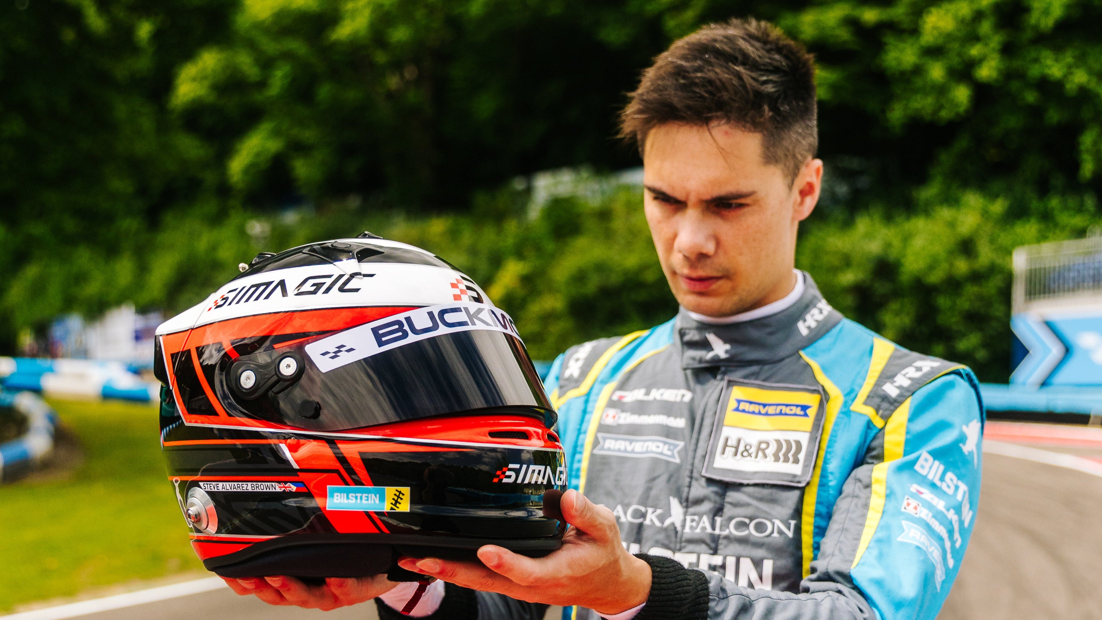 Racing driver (Super GT) holding a helmet with a visor sticker on a racetrack with greenery in the background